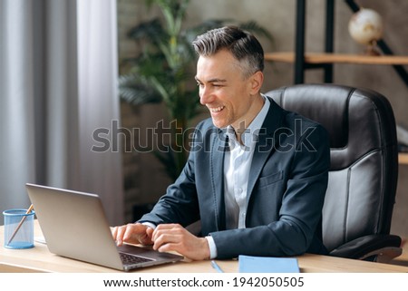 Influential successful Caucasian adult businessman or manager in a formal suit uses a laptop, communicates via video conferencing, online meetings, sits office, types on a keyboard, smiles friendly Royalty-Free Stock Photo #1942050505