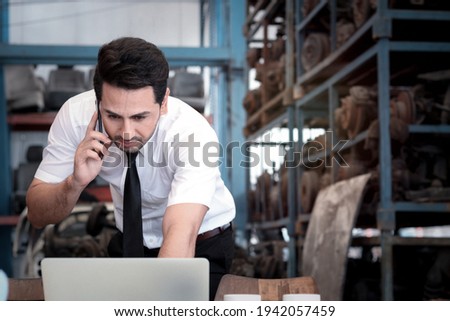 Portrait of smiling businessman seller working with laptop computer and talking on mobile phone at auto spare parts store shop warehouse with many second hand engine parts at blurred background.