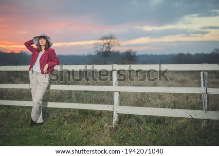 A young caucasian female in an elegant hat and pink jacket posing while leaning on a fence in a countryside field