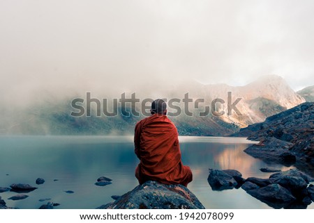 A Tibetan monk from back sitting on the stone near the water in the background of foggy mountains Royalty-Free Stock Photo #1942078099