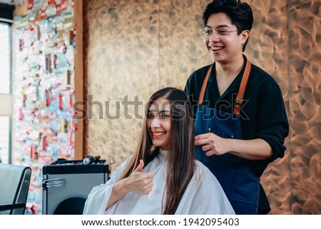Portrait of happy and cheerful young female customer showing thumbsup sign with male hairdresser wearing apron styling hair