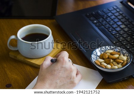 A person writing in a notebook while working on a laptop and drinking coffee