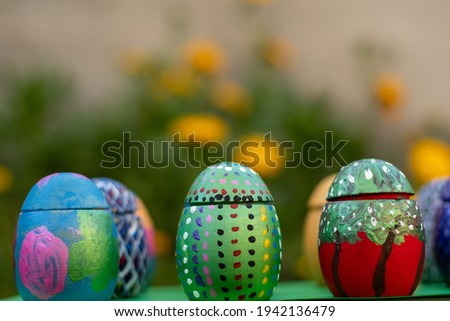 Hand-made Painted Wooden Easter eggs on a green pedastal in front of yellow and orange spring flowers.  There is a soft bokeh effect.