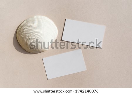 Still life scene with white big shell on beige background in sunlight and blank business, greeting card, invitation mockup. Long harsh shadows. Flat lay, top view.
