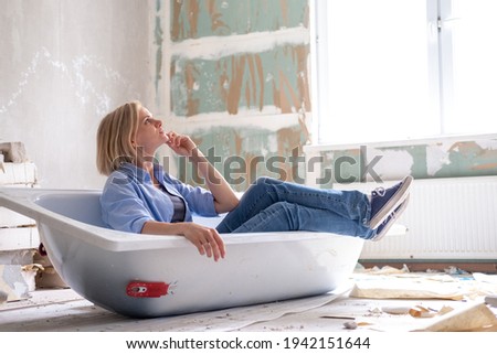 Renovation apartment. Creative story young dreaming plan woman sits in bathtub in the middle of the room. Empty walls, repairs house with their own hands. Royalty-Free Stock Photo #1942151644