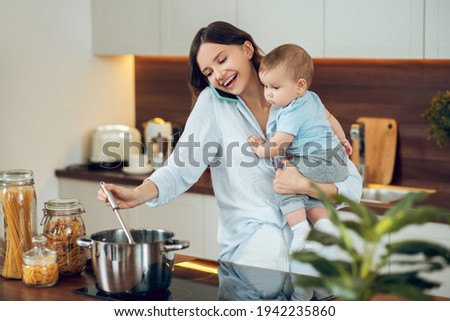 Cheerful woman with child talking on smartphone cooking Royalty-Free Stock Photo #1942235860