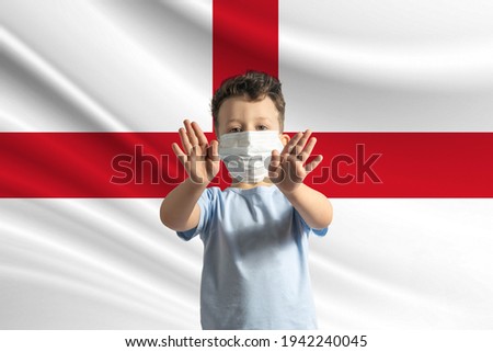 Little white boy in a protective mask on the background of the flag of England. Makes a stop sign with his hands, stay at home England.