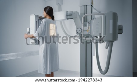 Hospital Radiology Room: Beautiful Multiethnic Woman Standing in Medical Gown in the X-Ray Machine. Adult Female Undergoes Healthcare Exam and is Scanning Chest, Heart, Lungs in Modern Clinic Office. Royalty-Free Stock Photo #1942278343