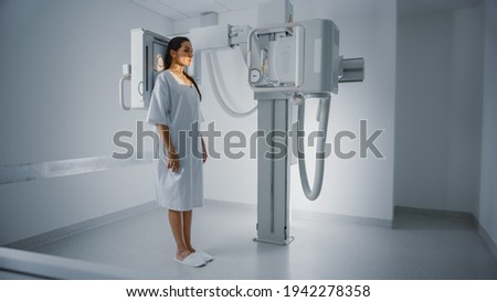 Hospital Radiology Room: Beautiful Multiethnic Woman Standing in Medical Gown in the X-Ray Machine. Adult Female Undergoes Healthcare Exam and is Scanning Chest, Heart, Lungs in Modern Clinic Office. Royalty-Free Stock Photo #1942278358