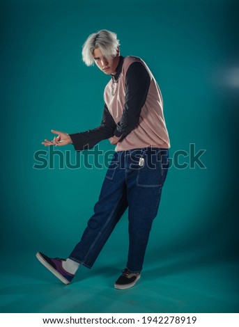 Young adult man in casual outfit wear looking at camera, holding skateboard near face and standing at studio on blue background