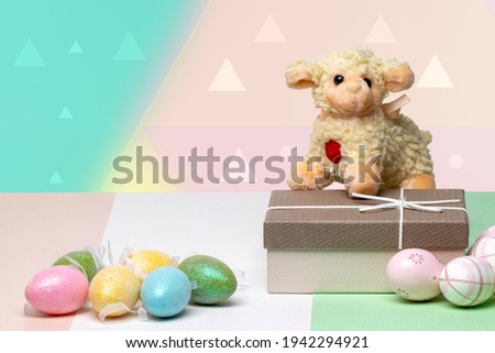 Happy Easter greeting card. Decorative composition of colorful eggs, a gift box with a easter lamb on it on a multicolour table in front of modern abstract pastel background. Copy space. Religion and 