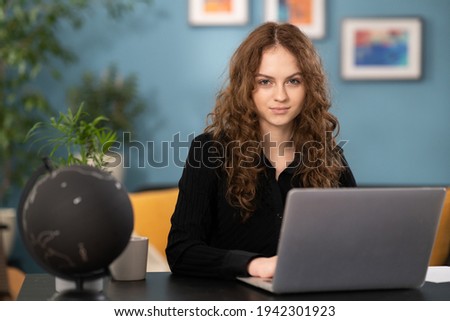 Female is using laptop, browsing websites and typing. Pretty student girl is studying remotely from her home. The girl is sitting at a desk in a cozy living room looking at the camera.