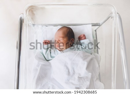 Newborn baby is sleeping in small transparent portable plastic bed. Baby first days of life is lying in a hospital crib after birth.
