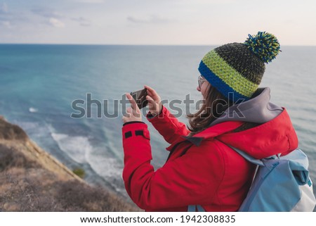 Young happy woman in hat and red jacket traveling taking photo on smartphone on rocky seashore