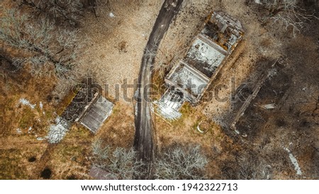 An aerial shot of a rural landscape with a dirt road across a meadow with an abandoned building with a collapsed roof and withered trees. Early spring landscape. Drone shot.