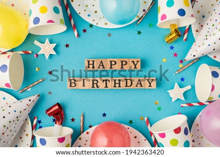 Top view photo of birthday composition wooden cubes labeled happy birthday in the middle polka dots paper cups and plates sequins balloons party accessories on isolated blue table background