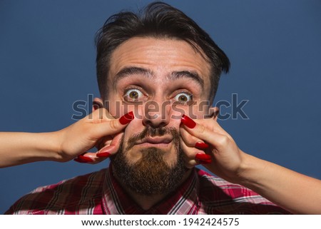 Patience. Half-length portrait of young handsome man with female hands punchning his face isolated on blue background. Concept of mood, psychology, feelings, funny meme emotion. Copy space for ad.