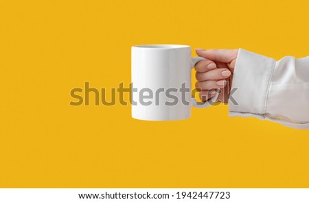 mockup ceramic white coffe cup or mug in female hands on yellow background with copy space. Blank template for your design, branding, business. Woman in shirt casual style. Real photo. Royalty-Free Stock Photo #1942447723
