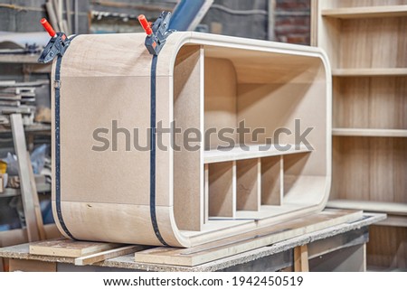 Making curved casing of new wooden cabinet with shelves using contemporary band clamp on work bench in carpentry workshop Royalty-Free Stock Photo #1942450519