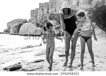 Mom and two little daughters walk along the seashore in swimsuits, looking at the sand. Family vacation at the sea.