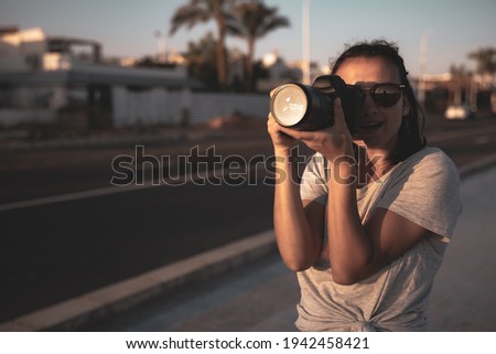 Young woman in summer in a T-shirt with a professional camera makes a photo at sunset in summer. Tourism concept.