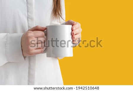 mockup ceramic white coffe cup or mug in female hands on yellow background with copy space. Blank template for your design, branding, business. Woman in shirt casual style. Real photo.