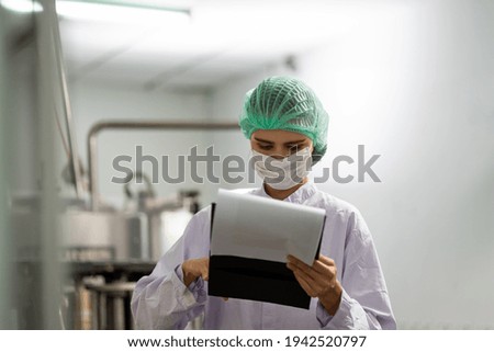 Female employee worker wearing mask, hairnet and using clipboard checking product of Basil seed with fruit on the conveyor belt in the beverage industry factory. Inspection quality control