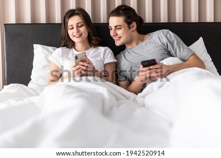 Portrait of jealous young man looking at his girlfriends mobile phone while sitting together in bed at home