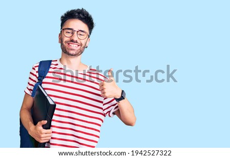 Young hispanic man wearing student backpack and glasses holding binder smiling happy and positive, thumb up doing excellent and approval sign 