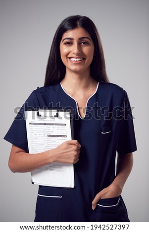 Studio Portrait Of Smiling Female Nurse With Clipboard Wearing Uniform Against Plain Background