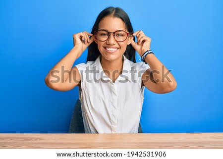 Beautiful hispanic woman wearing casual clothes sitting on the table smiling pulling ears with fingers, funny gesture. audition problem 