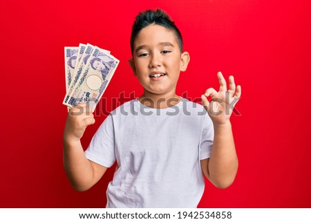 Little boy hispanic kid holding 5000 japanese yen banknotes doing ok sign with fingers, smiling friendly gesturing excellent symbol 