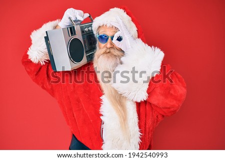 Old senior man wearing santa claus costume and boombox smiling happy doing ok sign with hand on eye looking through fingers 