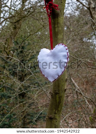 A home made heart in white material hanging from a tree with red ribbon in a wooded aera