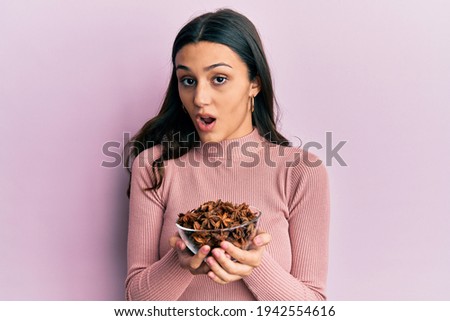 Young hispanic woman holding bowl of star anise in shock face, looking skeptical and sarcastic, surprised with open mouth 