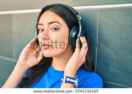 Young hispanic woman with serious expression listening to music using headphones at the city.