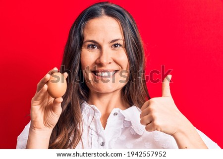 Beautiful brunette businesswoman holding bunch of dollars banknotes over red background smiling happy and positive, thumb up doing excellent and approval sign