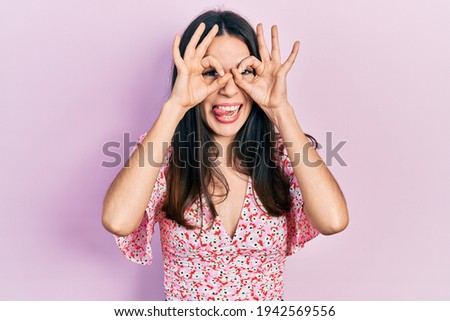 Young brunette woman wearing summer dress doing ok gesture like binoculars sticking tongue out, eyes looking through fingers. crazy expression. 