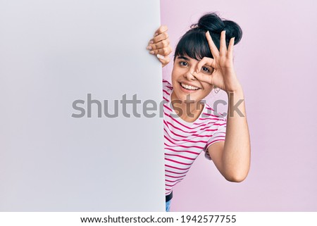 Young hispanic girl holding blank empty banner smiling happy doing ok sign with hand on eye looking through fingers 