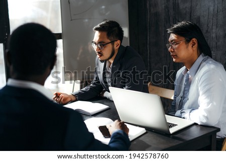 Serious men in formal suits are sitting at the table working at the computer and having a conversation