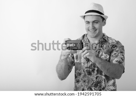 Studio shot of young happy muscular tourist man smiling while taking picture with mobile phone against white background