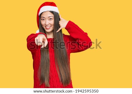 Young chinese woman wearing christmas hat smiling doing talking on the telephone gesture and pointing to you. call me. 