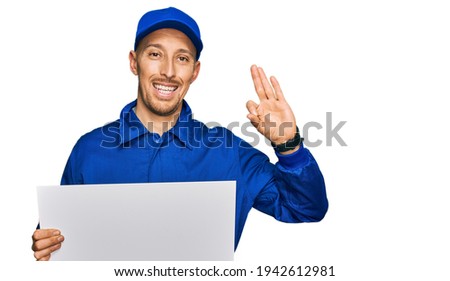 Bald man with beard wearing builder jumpsuit uniform holding empty banner doing ok sign with fingers, smiling friendly gesturing excellent symbol 