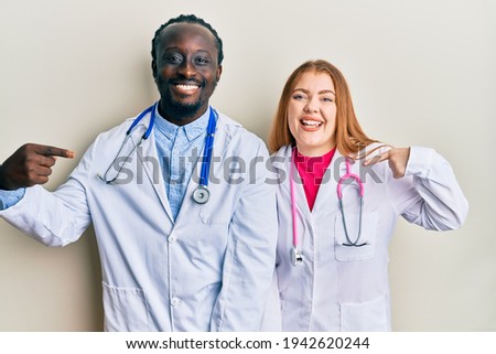 Young interracial couple wearing doctor uniform and stethoscope looking confident with smile on face, pointing oneself with fingers proud and happy. 