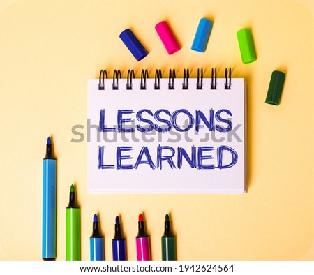 The words LESSONS LEARNED written in a white notebook on a beige background near multi-colored markers