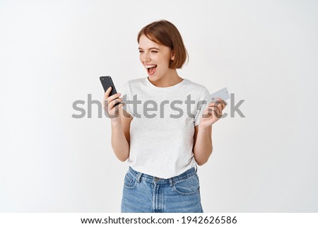 Happy smiling woman holding smartphone and plastic credit card, looking at mobile phone screen excited, making money online, standing against white background