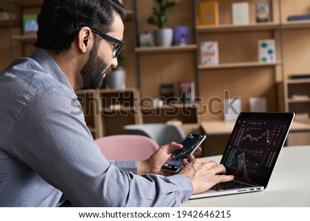 Indian latin business man trader investor analyst looking at financial stock market charts rate dynamic, trading data indexes growth using mobile phone and laptop for cryptocurrency digital analytics. Royalty-Free Stock Photo #1942646215