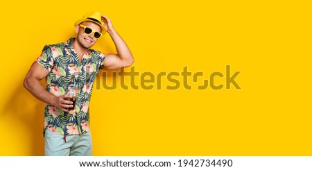 Handsome male in shirt, stylish sunglasses, cool yellow hat holding cocktail with straw posing in studio over bright yellow background
