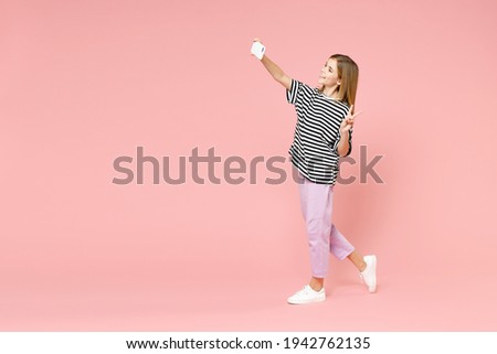 Full length little blonde kid girl 12-13 years old in striped oversized t-shirt doing selfie shot on mobile phone walking isolated on pink background children portrait. Childhood lifestyle concept