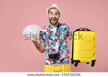 Surprised young traveler tourist man in summer clothes hat hold suitcase fan of cash money in dollar banknotes isolated on pink background. Passenger traveling on weekends. Air flight journey concept